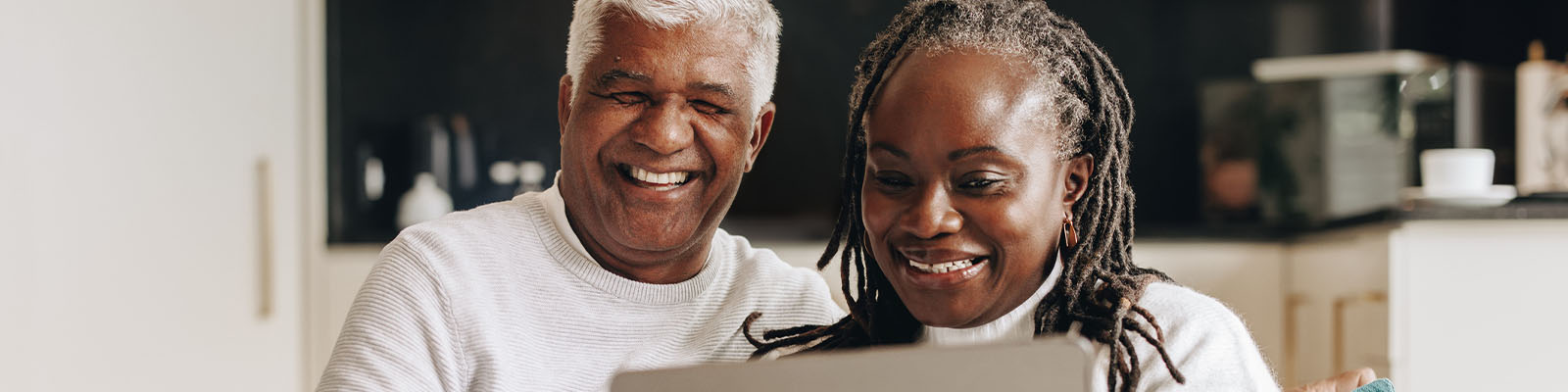 A senior man and senior woman sitting together on a couch looking down at a laptop while smiling