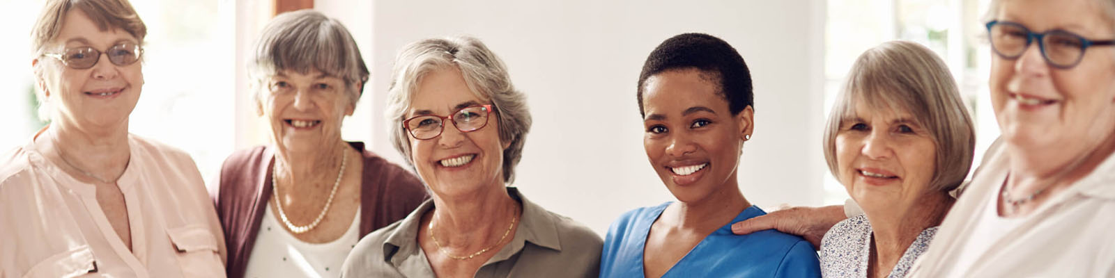A female senior nursing assistant in blue scrubs standing in the middle of senior women in a senior living facility