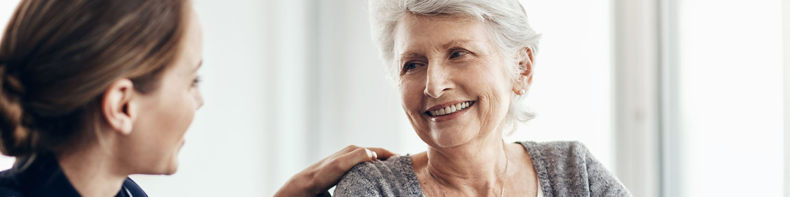 A senior woman sitting in a wheelchair looking at her nursing assistant while smiling