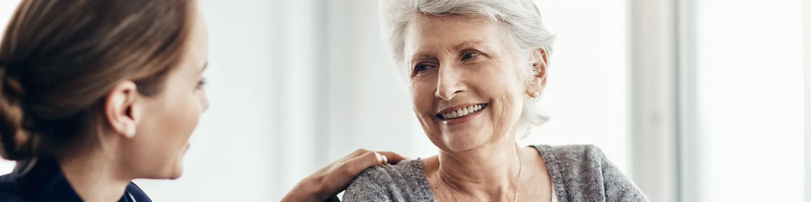 A senior woman sitting in a wheelchair looking at her nursing assistant while smiling