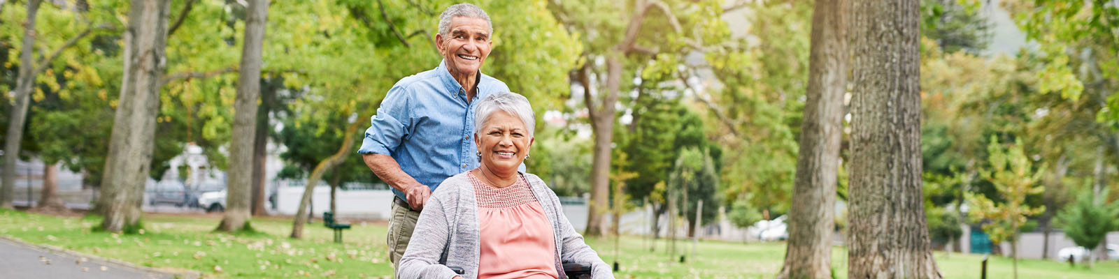 A senior man pushing his wife in a wheelchair outdoors on a walking path