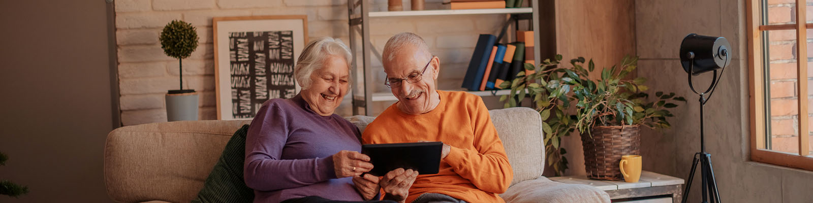 An older couple is sitting together on a grey couch. Both are holding an iPad, looking down at it and smiling.