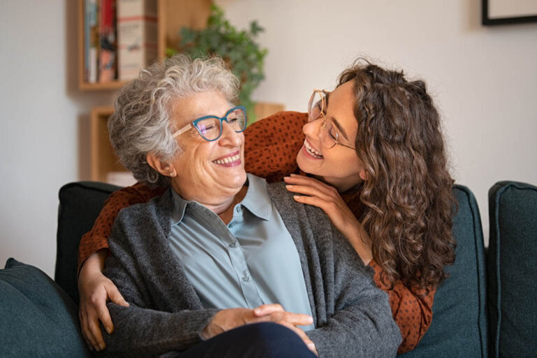 Female assisted living resident at Traditions of West Chester sitting on her couch as her daughter leans down to hug her