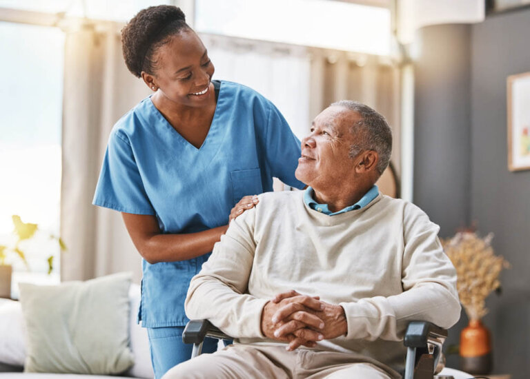 Male memory care resident sitting in a wheelchair in his senior apartment as his female caregiver leans down to check on him