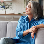 A senior woman wearing jeans and a blue shirt sitting on a grey couch while looking out a window with the Traditions Management logo in the top left corner of the image